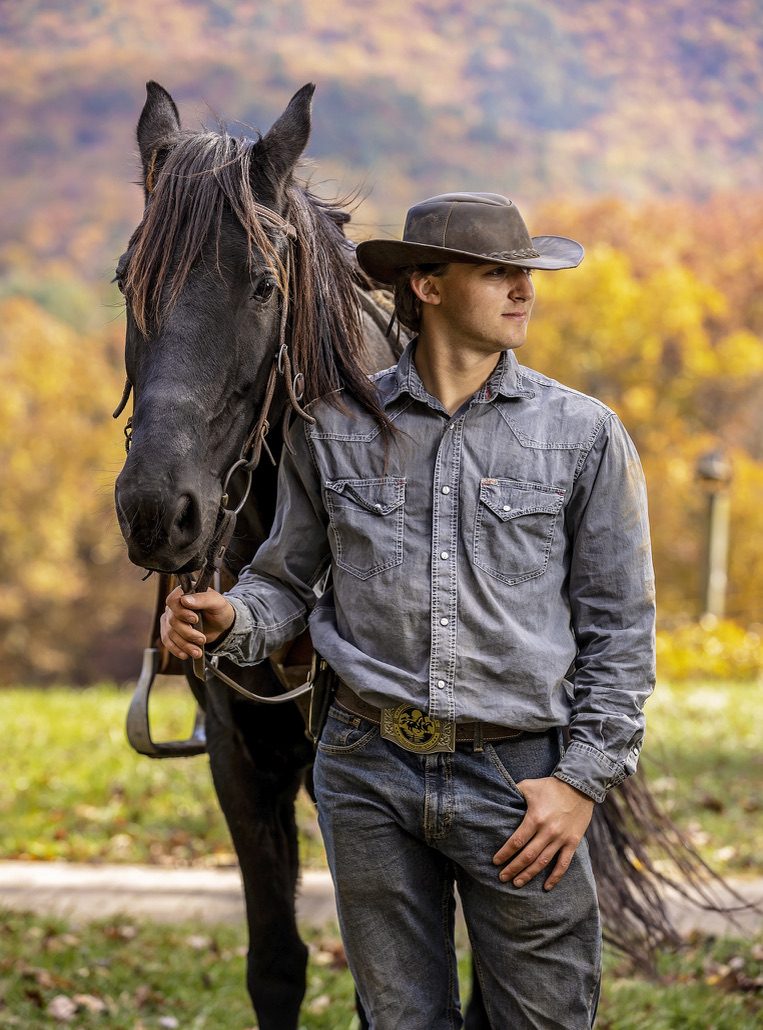 Young rider in a hat leading a black Friesian horse in fall light