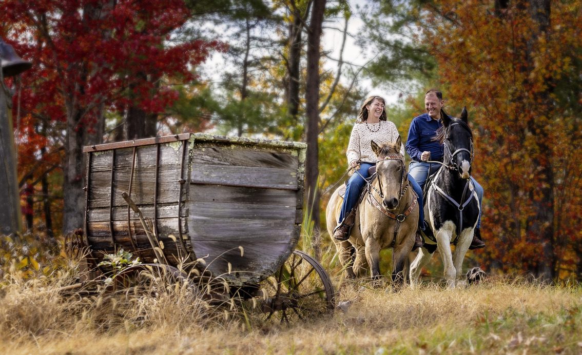 Two riders laughing on a trail ride past an antique wagon in fall color